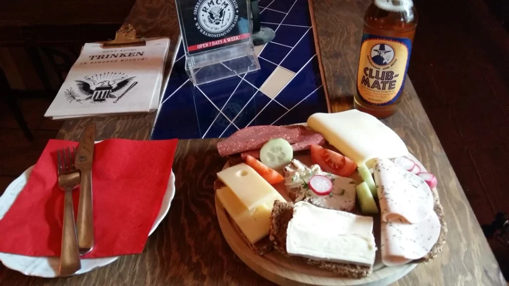 Savory meat and cheese platter with fresh vegetables, along with a bottle of Club Mate soda, served on a rustic wooden table at a cozy pub or restaurant.