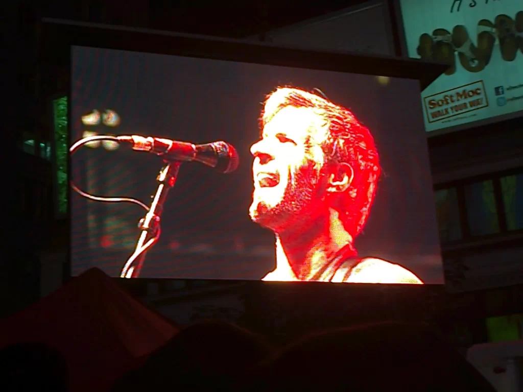 A screen showing Bad Religion's bassist Jay Bentley, a middle aged man but with longer blonde hair, also singing into a microphone.