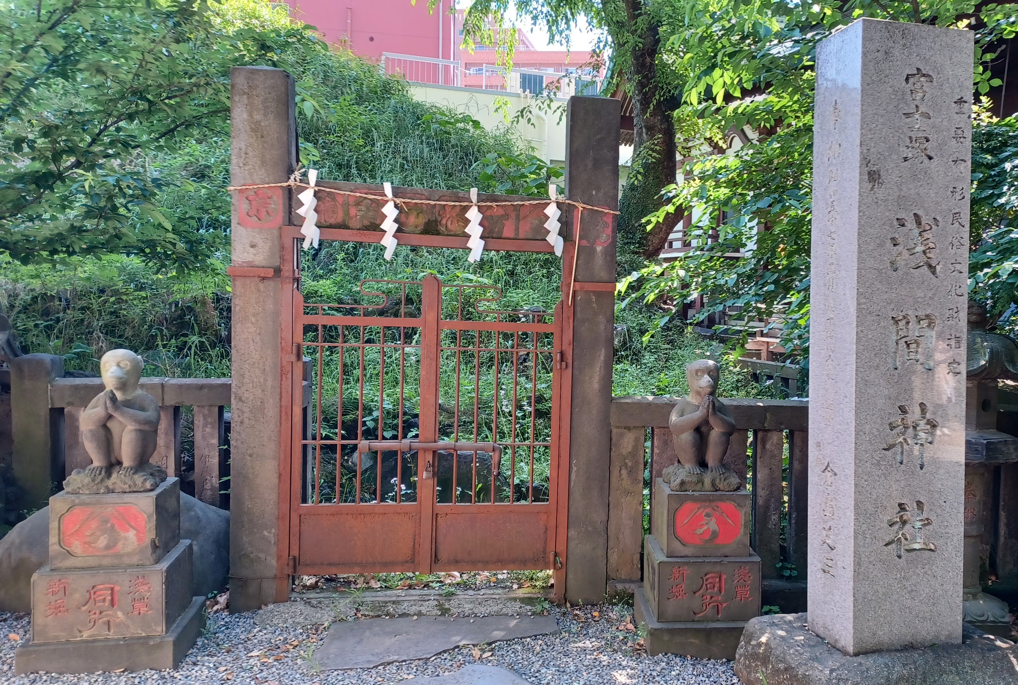 A brown gate with a stone monkey on the left and right side, who are guarding the miniature Mount Fuji. There's a high stone with Japanese symbols next to the monkey on the right side.