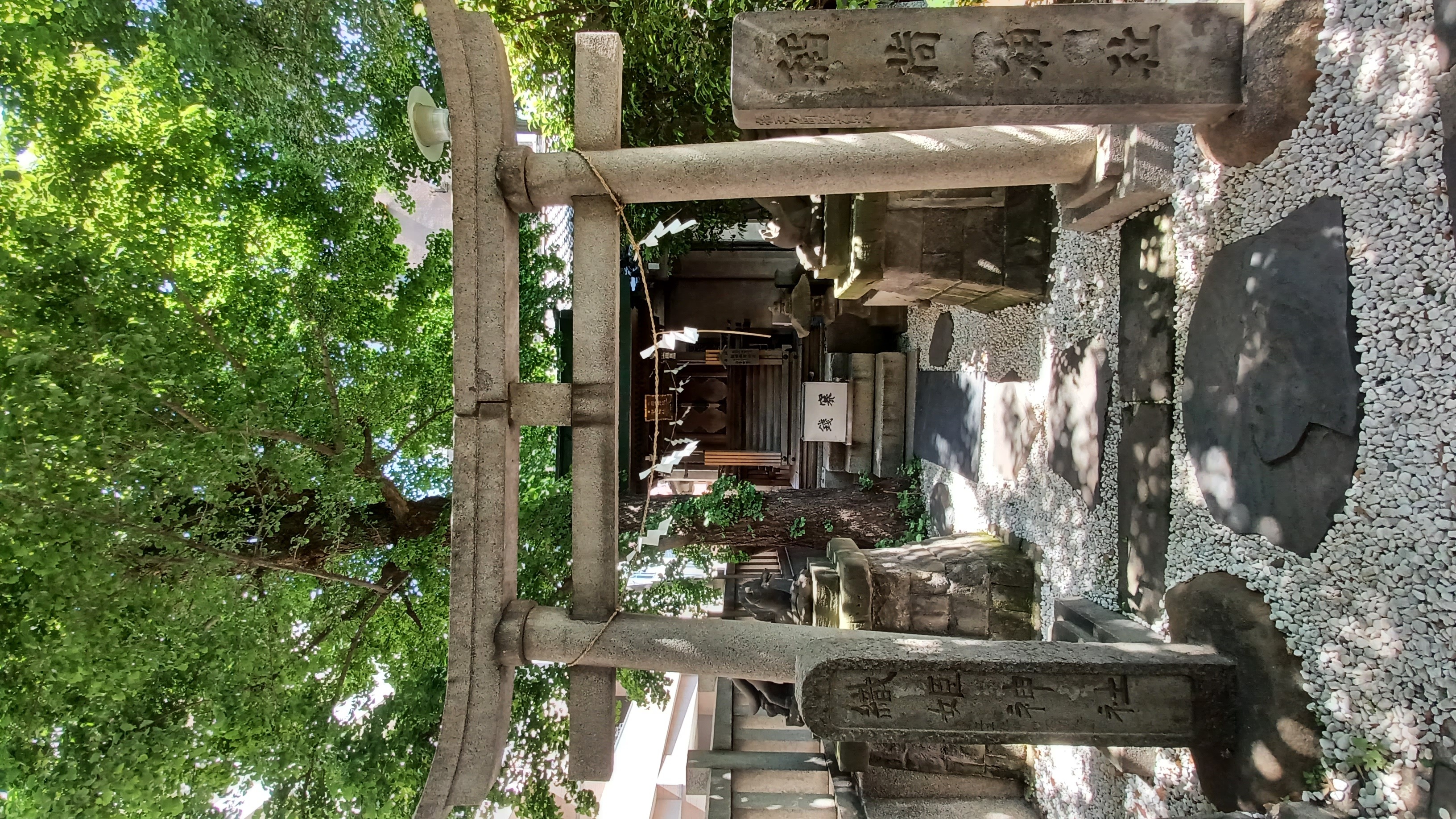 A grey torii (Japanese-style) gate standing in front of a sub-shrine building. There are two high stones with Japanese symbols on them next to the gate.