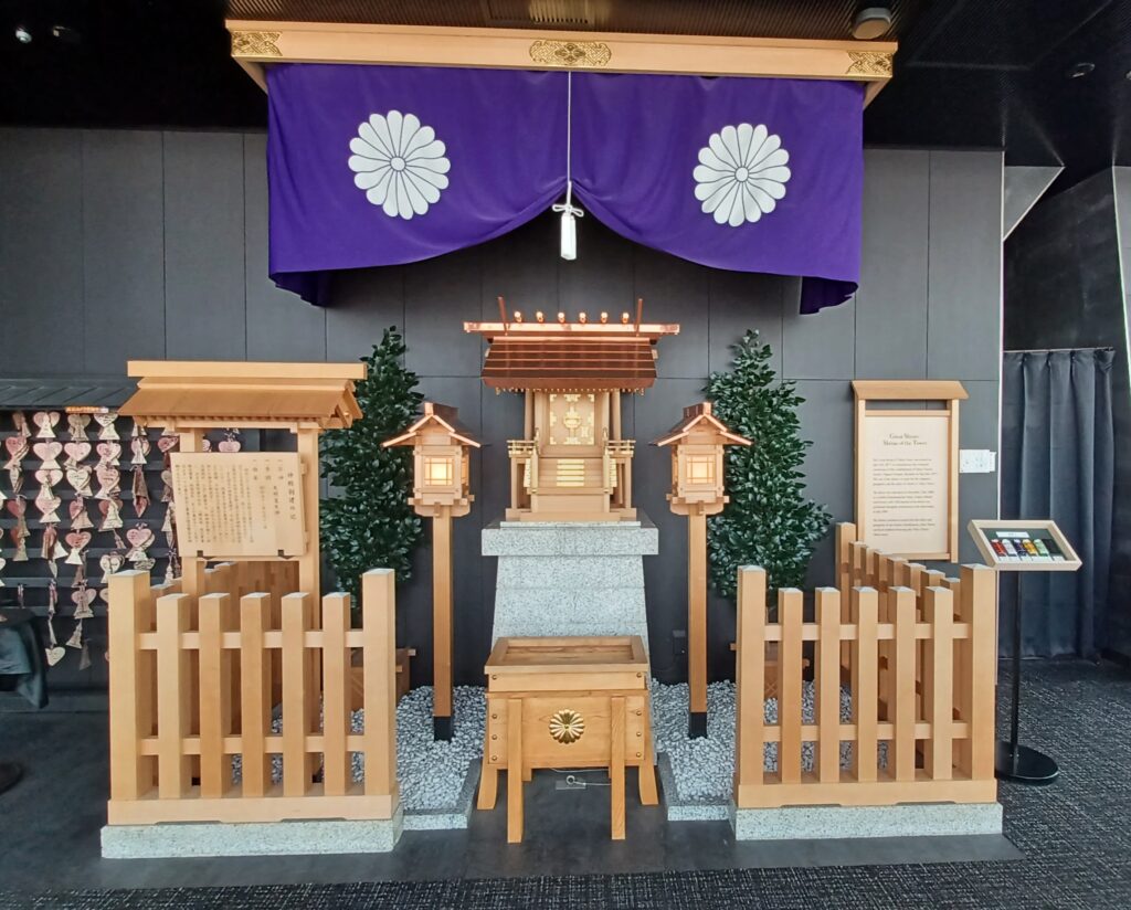 A small light wooden Japanese-style shrine with a purple banner above it. There's a white flower on each side and a thin rope in the middle.