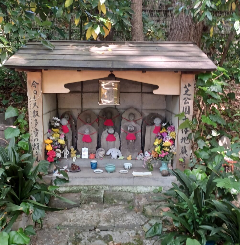 2 rows of the Jizo statues who were red bibs. There are flower bouquets on each side and, some little bowls, and stuffed animals in front of the first row of Jizo.