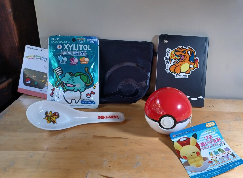 Pokémon merch: White soup spoon, black pouch, red/white Pokéball, Charizard sticker on a little black book, and a blue/white bag of sweets with Bulbasaur holding a toothbrush. 