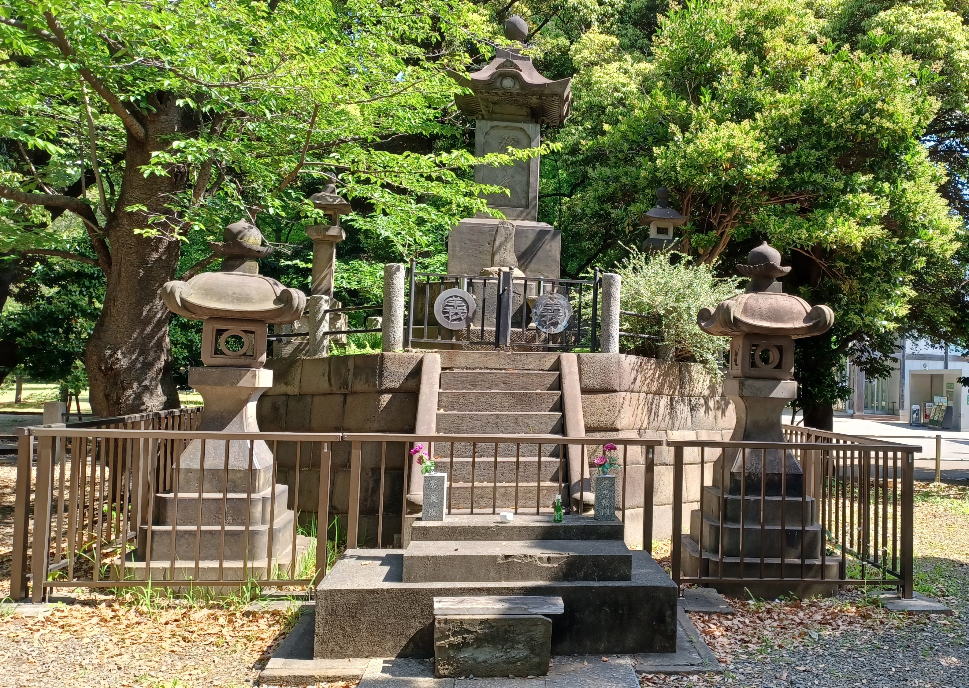 A stone memorial site, with a few stairs leading up to a large gated stone lantern. There are smaller stone lanterns around it and trees in the background. There's a wider gate protecting the site in the front. 4 stones squares are in front of the gate and flowers in a vase on the left and right side.