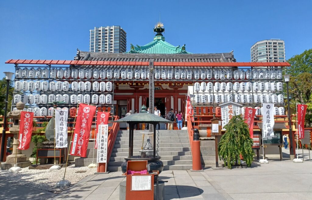 A temple hall with a turquoise roof, stairs leading up to the inside hall, and red and white flags in the front. There are also many white canisters with Japanese symbols written on them.