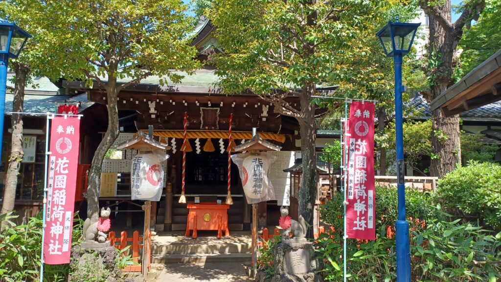 A dark brown prayer house  with two red/white ropes hanging from the ceiling. There's a small orange table in the front and two red flags with Japanese symbols on the left and right side (in front of the shrine).