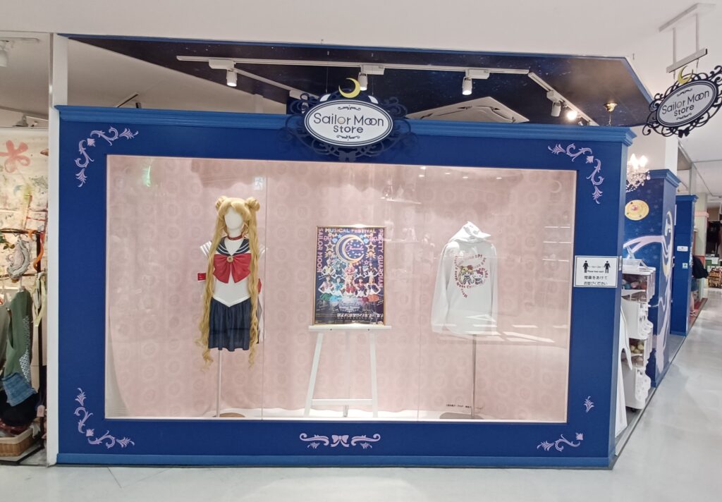 A store window with a dark blue frame around it, showing a Sailor Moon costume, poster, and a white hoodie with Sailor Moon on it.