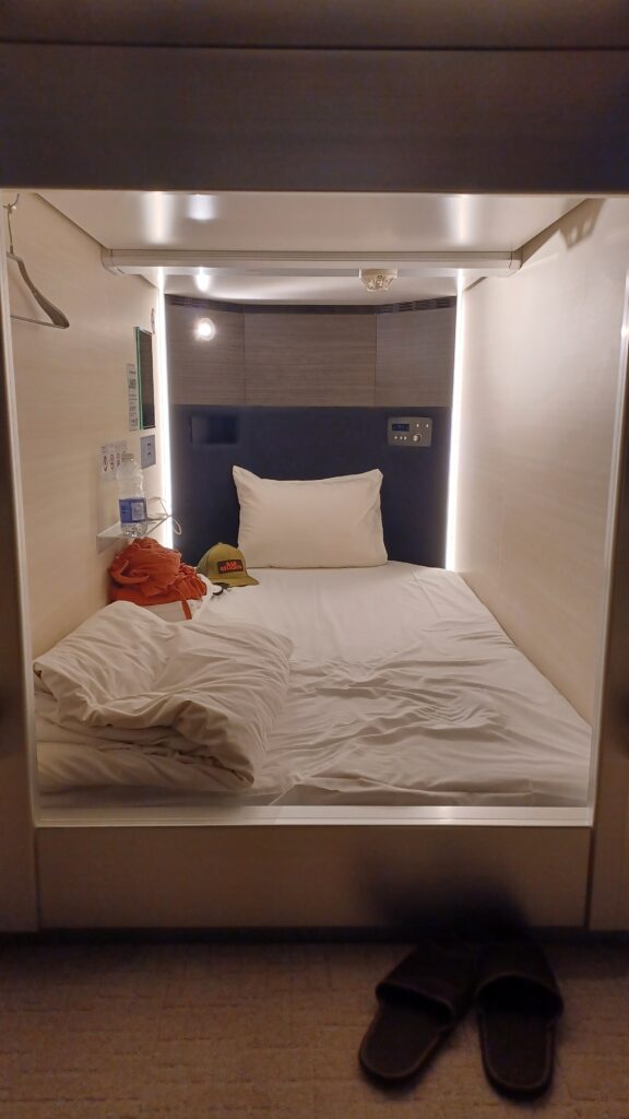A simple, but comfortable bed inside the capsule, with dark brown slippers in front of the capsule.