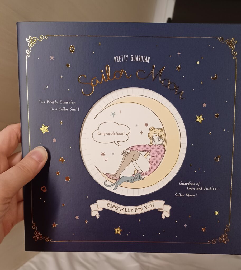 A dark blue green card with Sailor Moon and her cat sitting on a crescent moon. She says "Congratulations!"  and there are some stars as well, so it looks like a night sky!