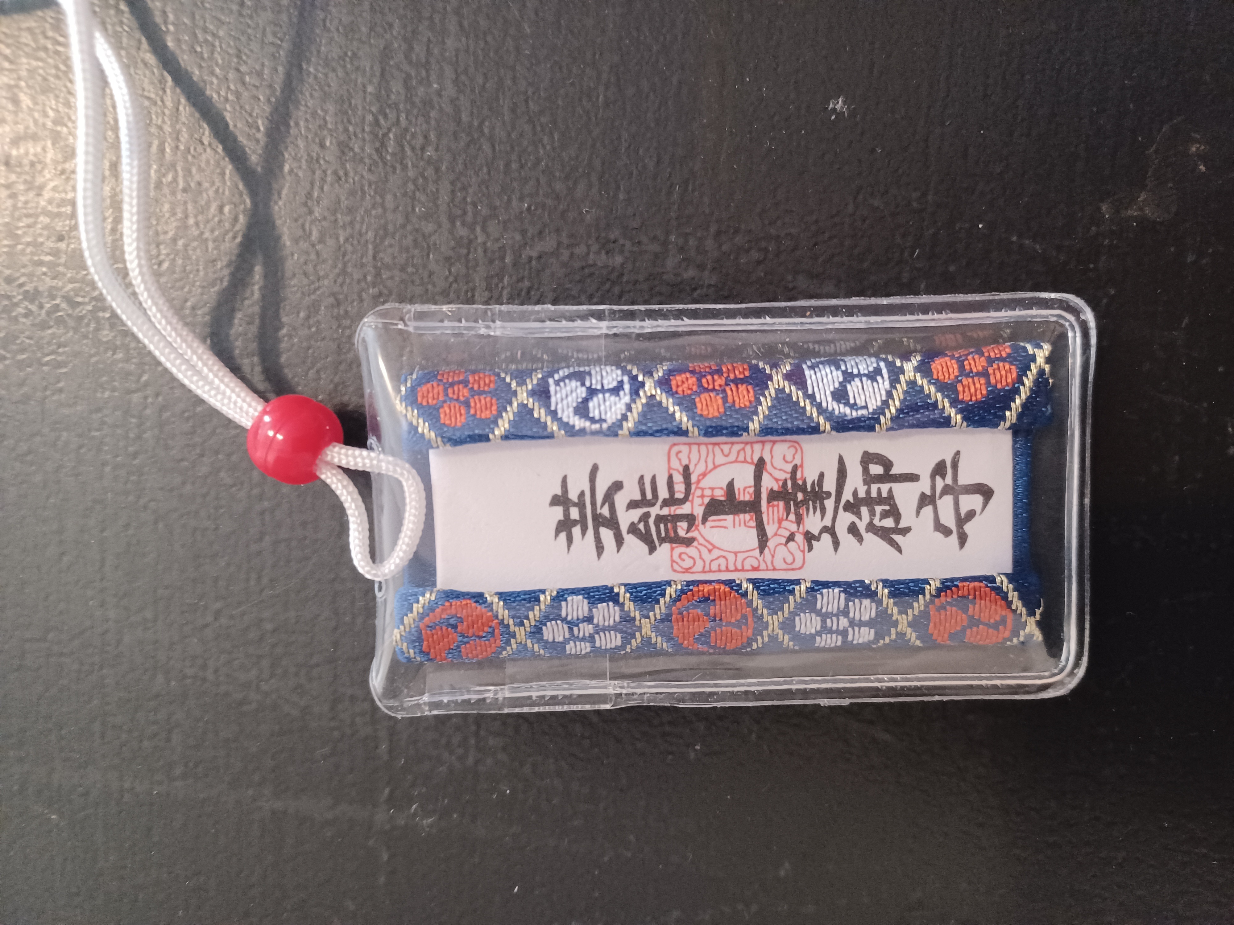 A blue/red/white lucky charm with Japanese symbols in the middle, and a white thread and red accessory at the top.