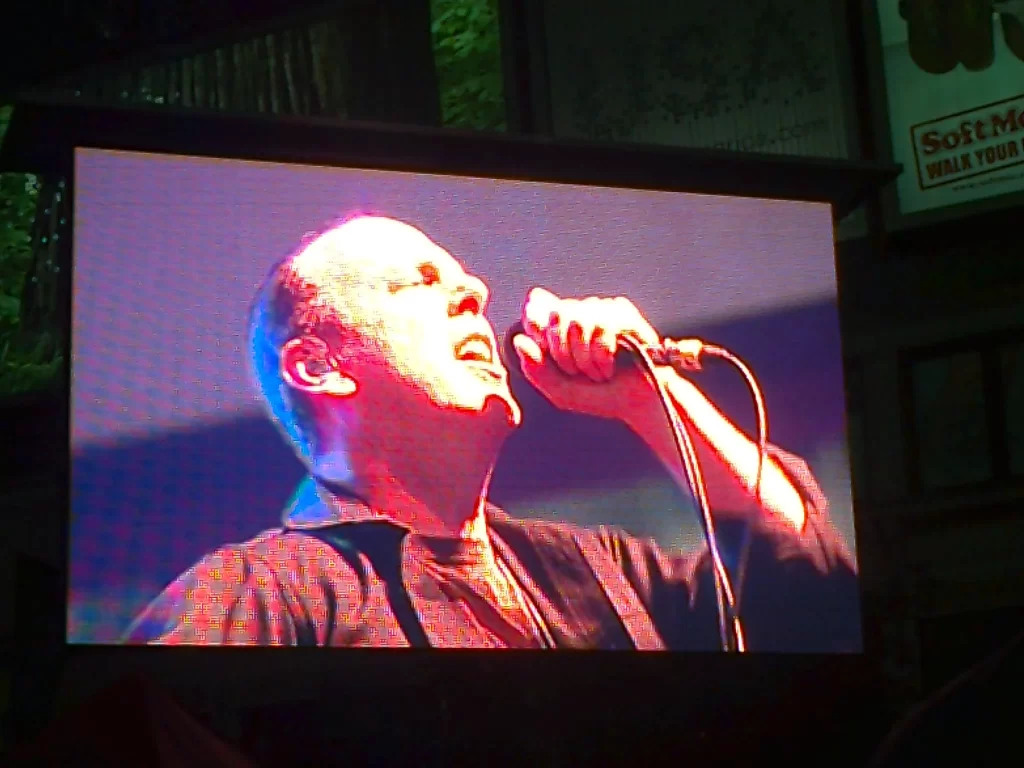 A screen showing Greg Graffin, the middle aged singer of Bad Religion with very short dark hair, who's singing into a microphone.