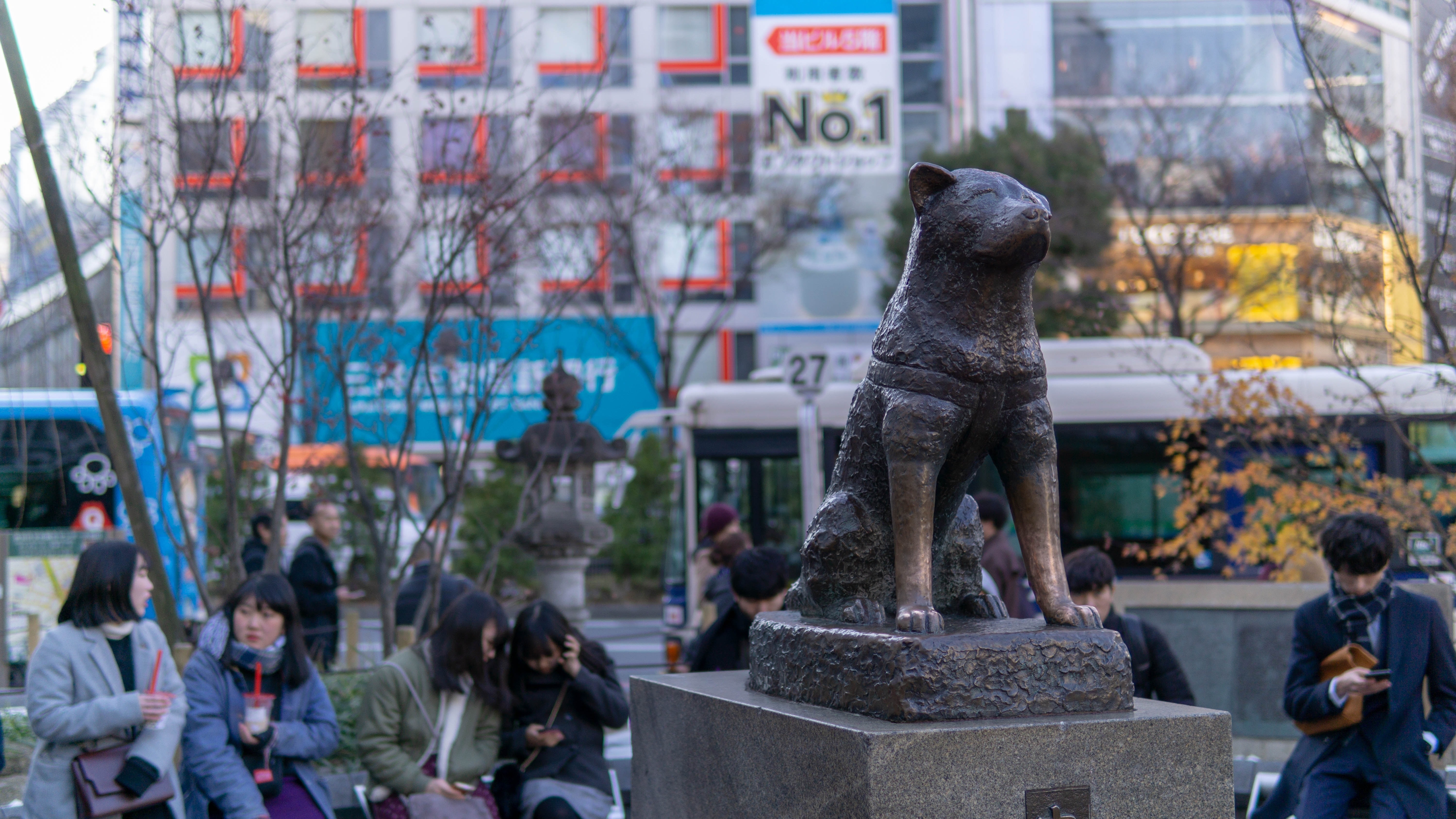 A dark brown dog statue sitting on a podium with people in the background.