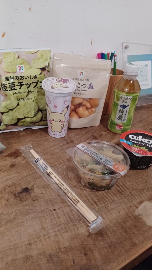 A selection of convenience store foods and drinks: Edamame chips, rice crackers, green tea in a bottle, strawberry yoghurt, veggie bowl, Pikachu-themed chocolates, and wooden chopsticks.