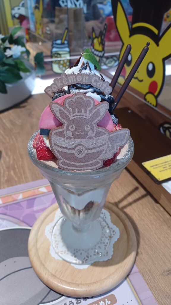 A large ice cream parfait that has a Pokémon Café logo in the front, with Pikachu in a bowl. It's wearing a chef's hat, and a Pokéball and raspberries are behind it. There's also some whipped cream and two chocolate sticks in the back.