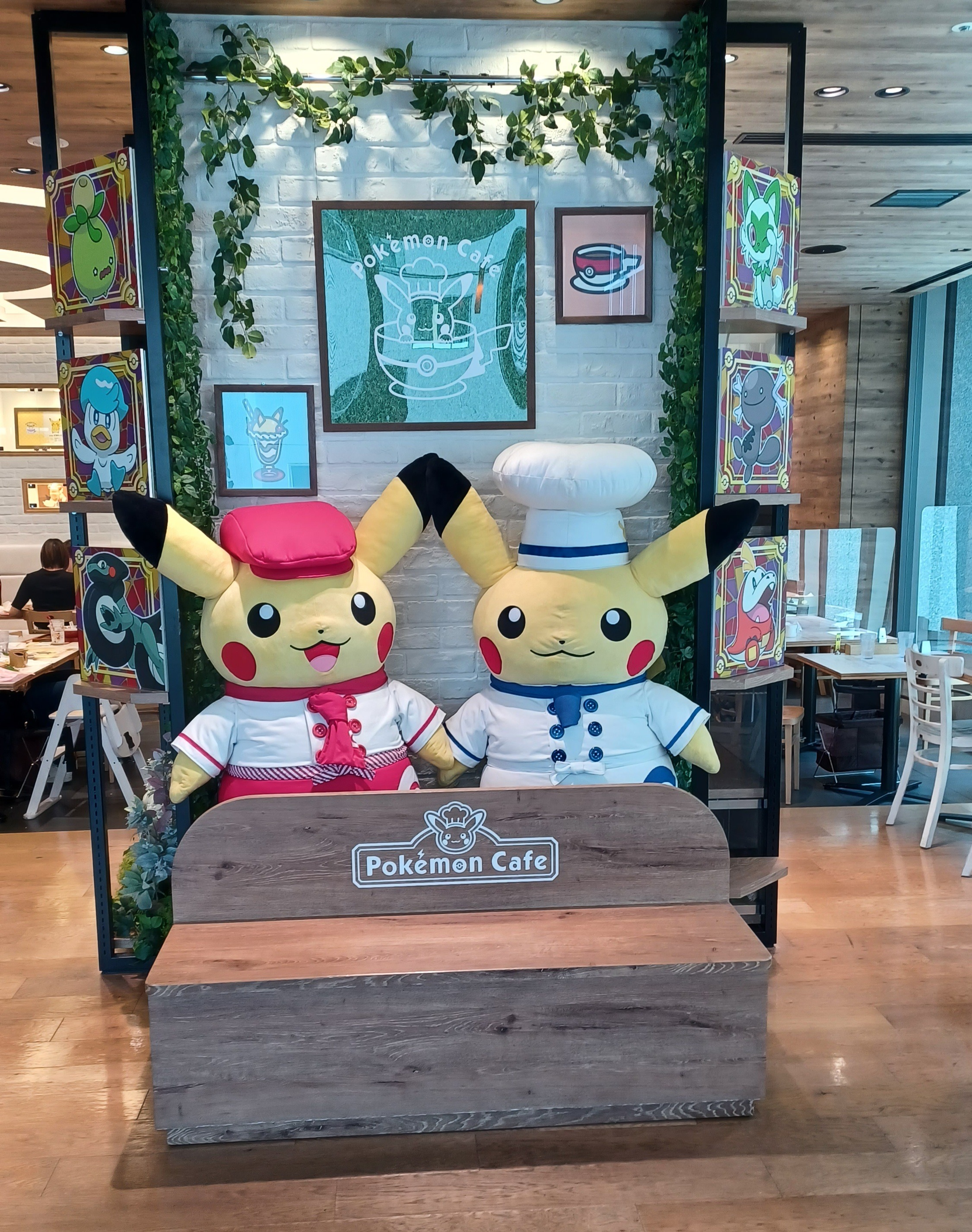 Two large Pikachu stuffed animals behind a Pokémon Café bench. The left one is dressed in a waiter's uniform, and the right one wears a chef's outfit. There are some picture of other Pokémon on the right side and an ivy plant in the back.