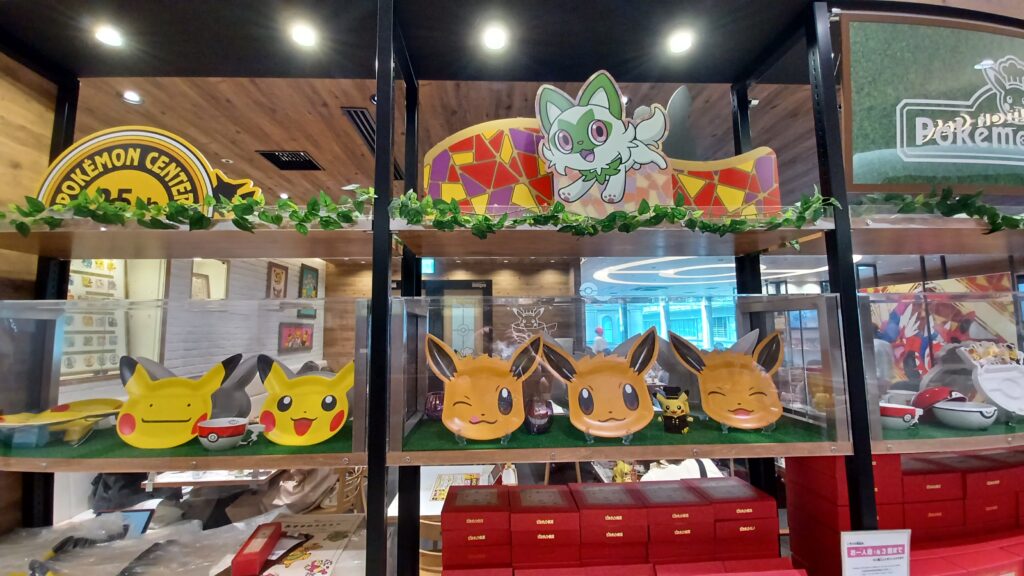 A shelf with a selection of Pikachu and Eevee plates and red/white Pokéball bowls for sale at the Pokémon Café. Underneath, there are red boxes with Pokémon soup spoons inside them.  