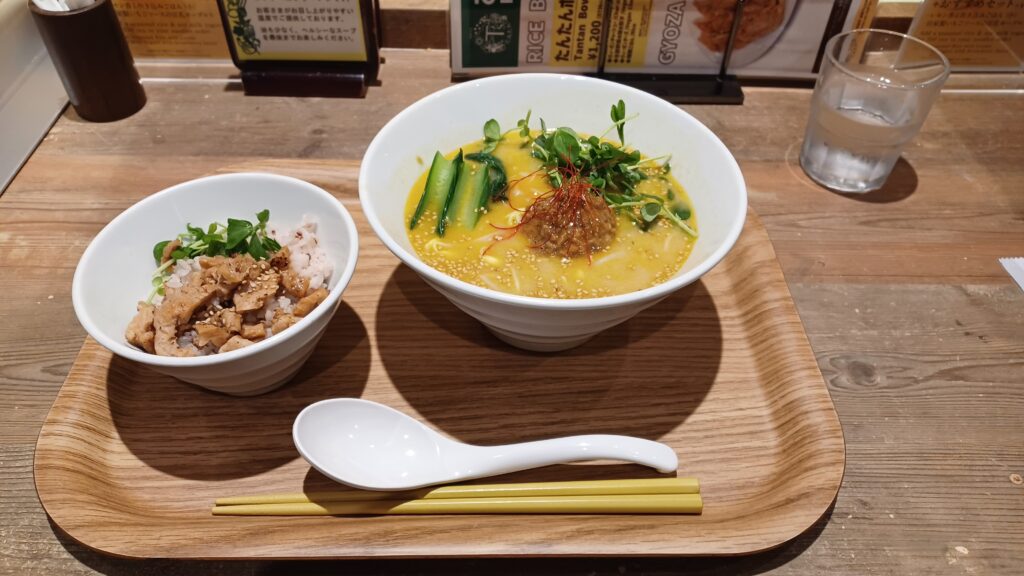 Right: A large ramen noodle bowl with yellow soup broth and green vegetables on top. Left: A small bowl with rice and soy meat on top. In the front, there's a white soup spoon and a pair of wooden chopsticks.