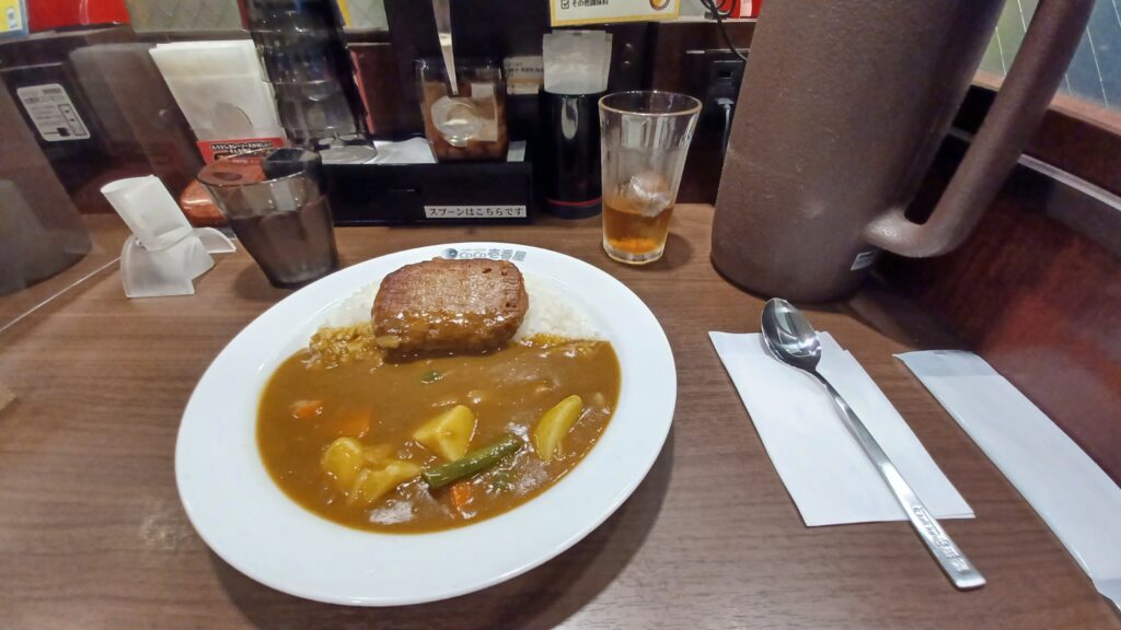 A large plate of Japanese curry, with rice and a soy meat burger on top. On the other side, there's a large portion of brown curry sauce with potatoes, carrots, and green beans.