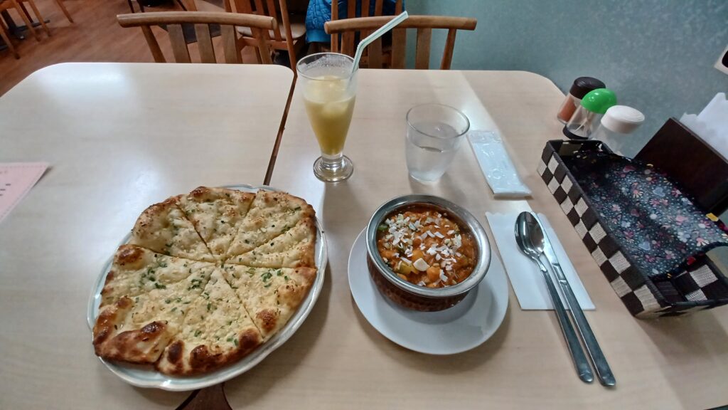 Garlic naan bread on the left, cut into pieces, Chana Masala on the right. Above these dishes there are a glass of Mango Lassi and water.