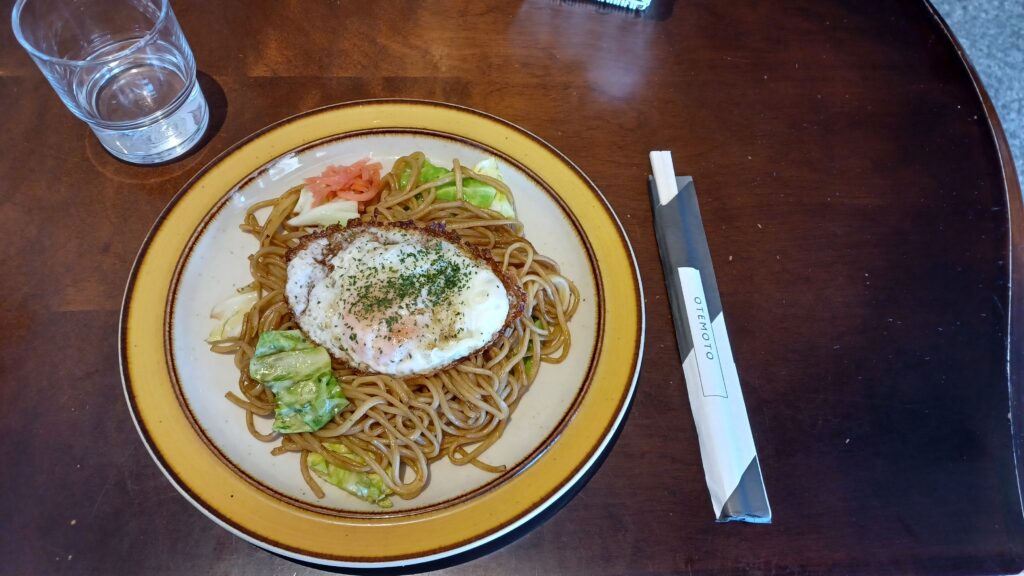 A plate of soba noodles with a few vegetables and a fried egg on top. On the left there is a glass of water.