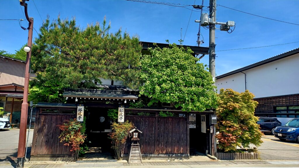 A dark brown wooden Japanese-style house with plenty of green plants on its roof. 