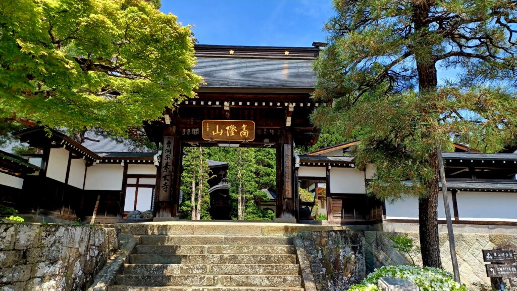 A dark brown temple gate entrance with a sign of golden Japanese symbols in the top middle part. A few trees are standing on the left and right side of the gate.
