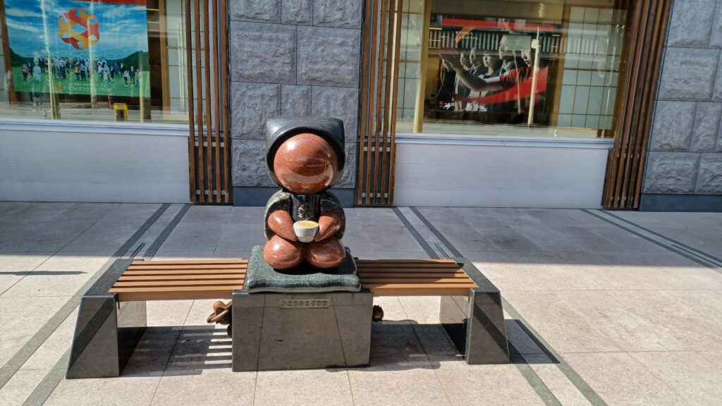 A red faceless Sarubobo statue sitting on a light wooden bench in front of a store window. It wears a black hat and jacket and holds a cup of coffee in its hands.