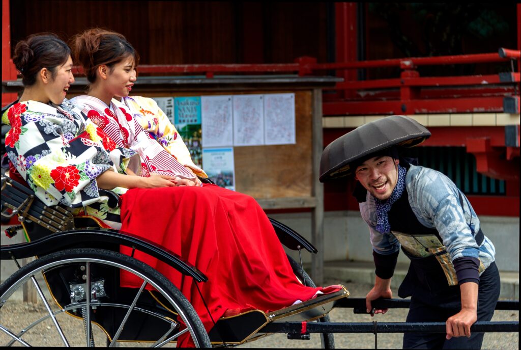 Two women in traditional Japanese dress are sitting in a carriage, with their legs being covered by a red blanket for comfort. A man, also dressed in traditional Japanese clothing, is wearing a black hat and will start pulling the carriage soon. All 3 of them are smiling!