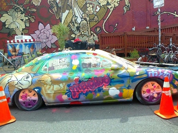A small car with a graffiti style exterior. Colour examples are pink, yellow, white, green, and blue. Green plants are growing out of its top area.