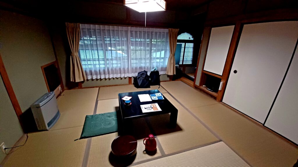 A simple, but bright room with light brown tatami flooring, AC/heater on the left, and a black short-legged table in the middle. There are some documents on the table, a red tea pot on the floor, and a green pillow to sit in front of the table. White, thin curtains cover the large windows, and there's a large closet with two sliding doors on the right hand side. 