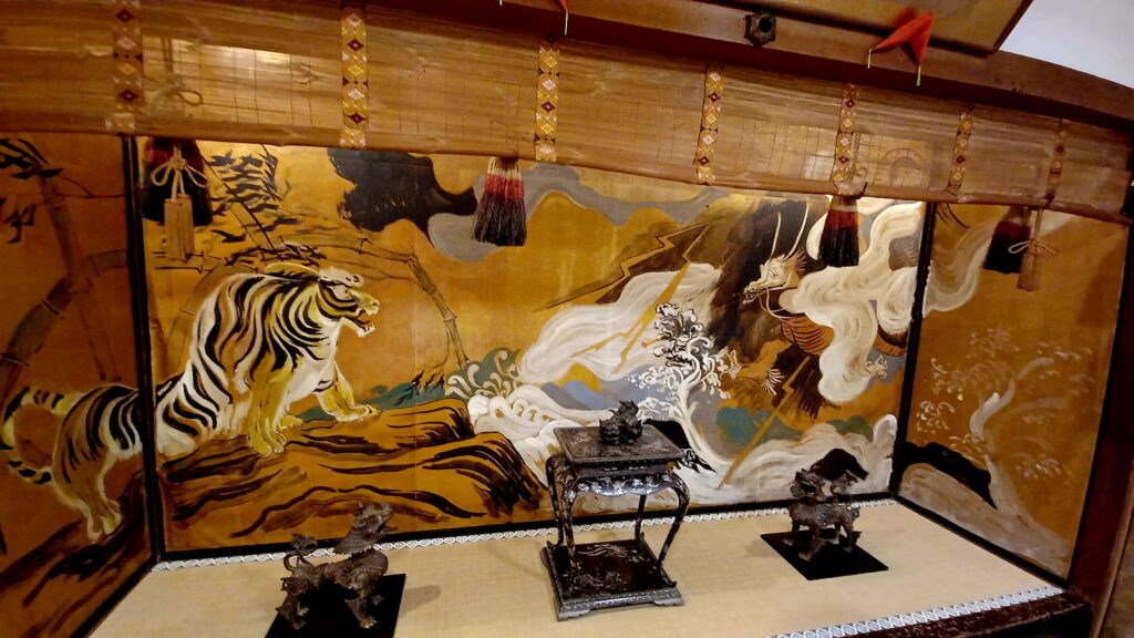 A colourful painting with a tiger standing on a cliff on the left side and white dragon on the right side. Both are separated by gorge filled with white/blue water. It also has bamboo stalks on the left side, and a mostly orange background. Two black dog-like statues sit on the left and right (in front of the painting canvas) and a black table in the middle. 