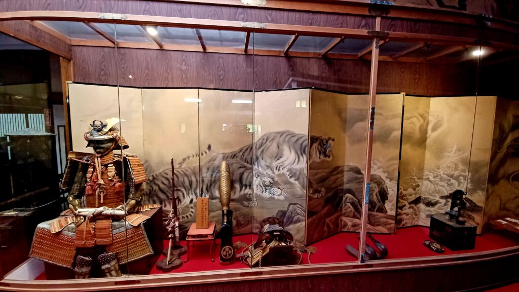 A black and white painting of two tigers next to each other on the left side, and ocean waves on the right side. There are clouds in the sky as well. In front of the painting, there's a large Samurai statue, two weapons, a Samurai helmet, and a black box with a small statue on top (left to right).
