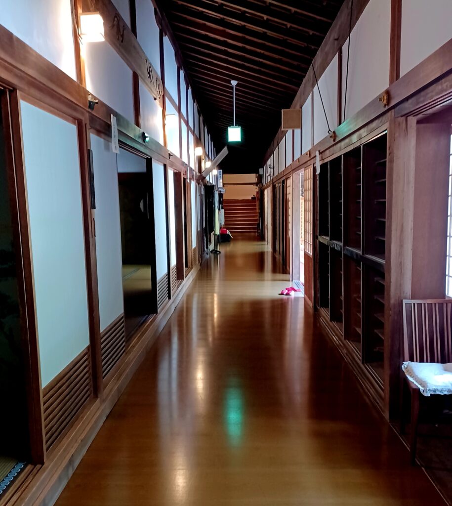 A long hallway with brown wooden flooring, and several white/dark brown sliding doors on the left. On the right, there's a dark brown closet with many shoe/slipper compartments.