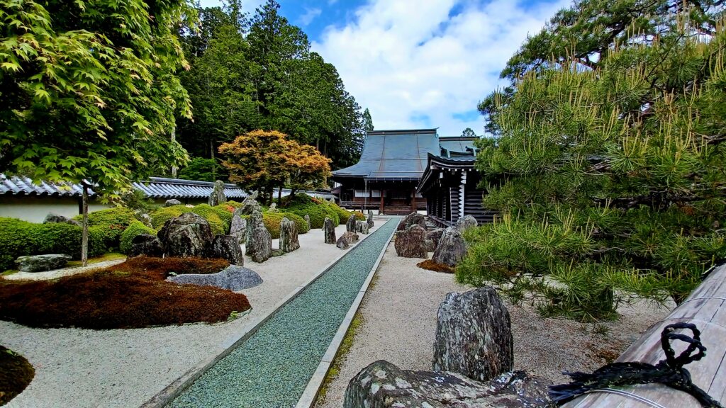 A large garden area with plenty of vertical rocks standing on the ground, and several green trees on the left and right side. In the middle, there's a pathway filled with little rocks that leads to a Buddhist worship building. 