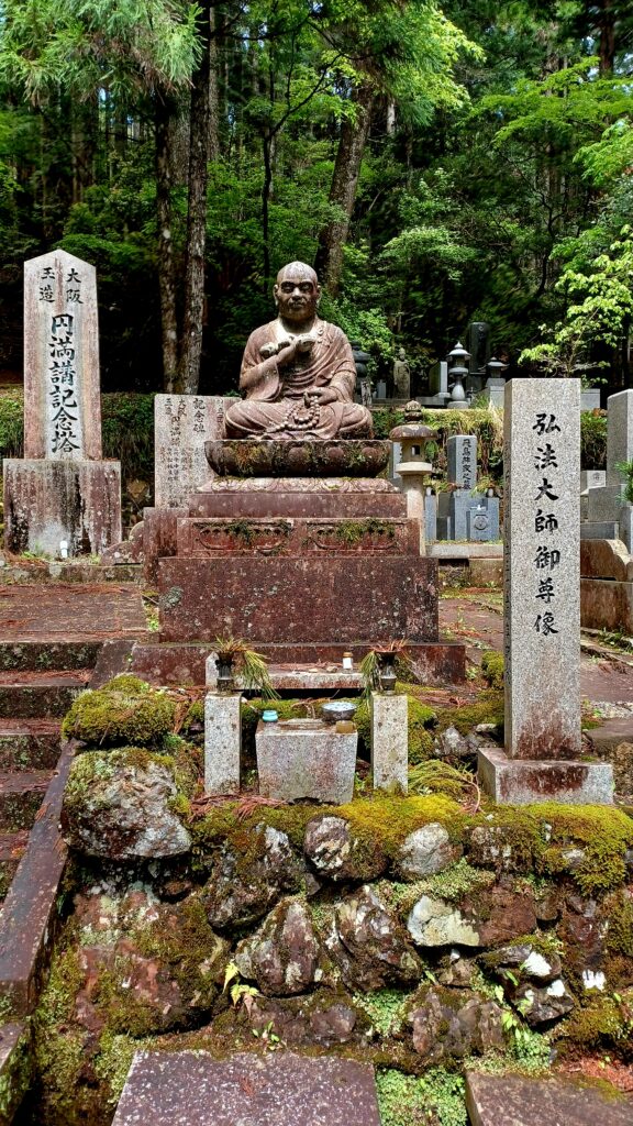 Several tombstones with a Buddhist figure (monk?) sitting on a stone chair in the middle of the picture. Trees are standing calmly in the background.
