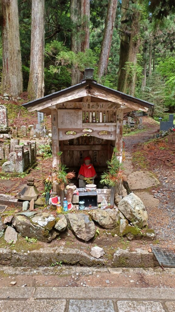 A tiny wooden hut with a single Buddhist Jizo  statue (it wears a red cape and red hat) sitting in front of it. There are larger rocks and a frew plants in front of it as well.