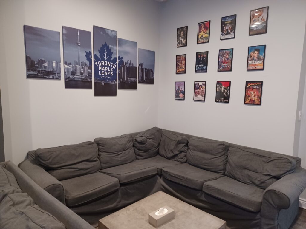 A large grey couch that goes around the corner with a light brown coffee table in the middle. On the one wall, there's a poster with the Toronto Maple Leafs team logo and some smaller framed movie posters on the other wall.
