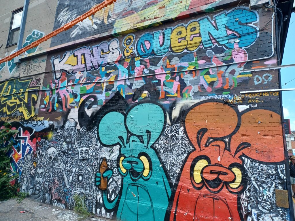 A colourful graffiti style artwork, with the title "Kings & Queens", and a bunch of skeleton figures. In the front, there's a blue and orange bunny. Their eyes are yellow with no pupils, and the blue bunny is holding a carrot.