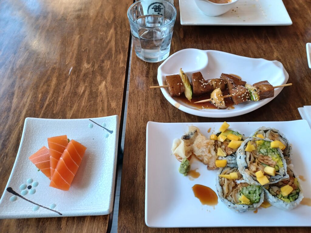 A plate of pink vegan salmon sashimi, soy beef skewers with vegetables, and large sushi rolls with mango pieces on top.