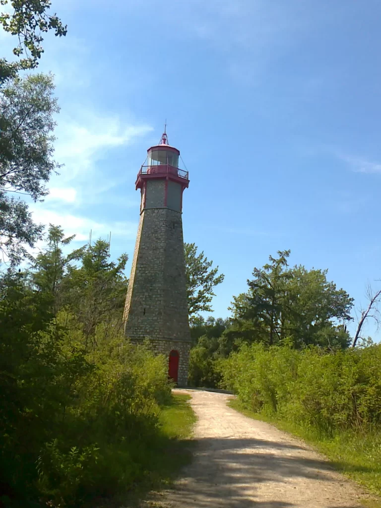 A lighthouse made out of grey cobblestones with a red door and red lookout area at the top. It's surrounded by green trees and bushes.