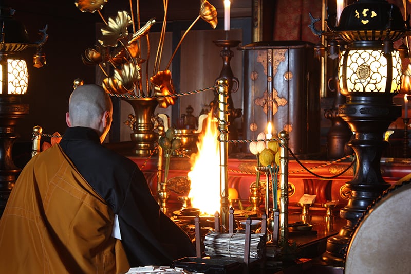A Buddhist monk is sitting in front of a small fire. He wears black and gold clothing and is surrounded by Buddhist treasure (for example, a bouquet of flowers or leaves (?) out of metal). Two large lamps are standing between the monk. There's candle light and incense as well. 
