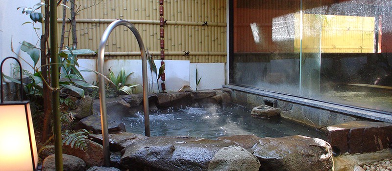 A small open air bath surrounded by large brown rocks and some bamboo plants. There's a silver handrail, so it's easier for guests to enter the bath.