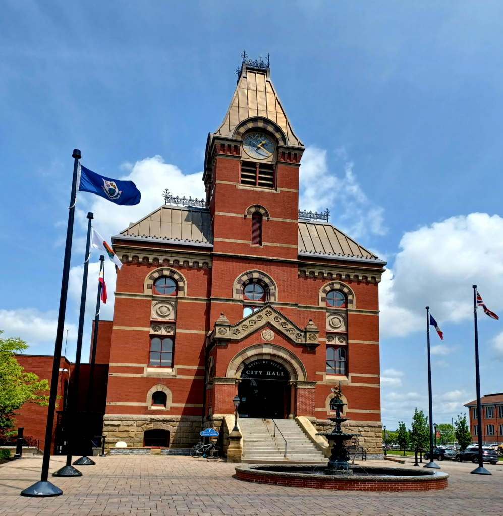 A high mostly red Victorian brick building with a triangular roof tower and large clock in the centre. There are flags on both sides of the building, e.g. the British and French flag. The sign says "City Hall", and if you go down the stairs, there's a large round fountain with a statue at the top.
