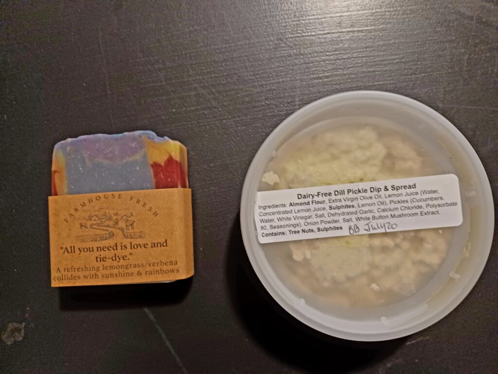 A rainbow coloured piece of soap and a little container of a dairy-free dill pickle dip and spread, showing its ingredients and best before date.