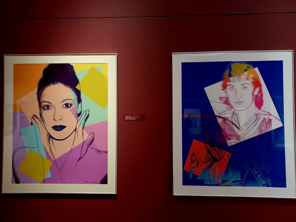 Two colourful paintings next to each other. The one of the left displays a woman with black hair and lips, holding her hands next to the face. She's surrounded by pink, orange, purple, green, and turquoise colour squares. The man on the right painting has red/orange/pink hair, and his face sits on top of a white square. The rest of the painting is dark blue with a little red square, that shows some letters.