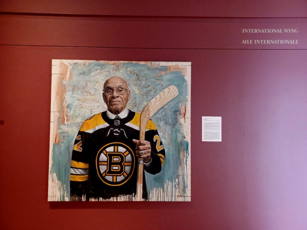 A painting of a bald Afro American man with glasses, who's wearing a shirt of the Boston Bruins ice hockey team and holding a light brown hockey stick in his hand.
