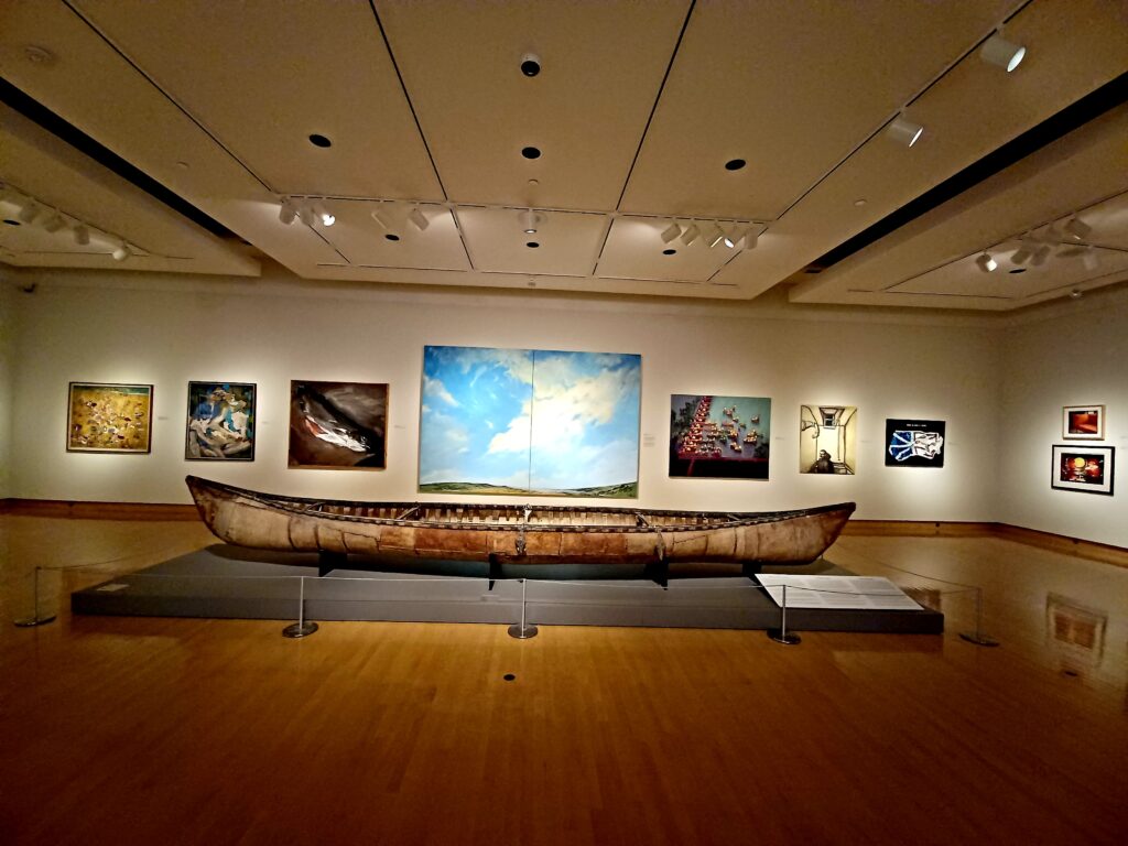 A large wodden canoe is sitting on a grey display area. Behind it, there are several paintings on the wall, with on showing large clouds in the sky.