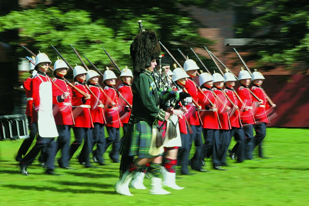 A military parade with most men dressed in a red jacket, white hats, and dark blue pants. They all carry a rifle on their shoulder and walk on grass. At the front, there's a man dressed in a Scottish artire (green kilt and jacket with a furry high black hat) playing bagpipes. The man next to him is wearing a red kilt and playing bagpipes as well. 
