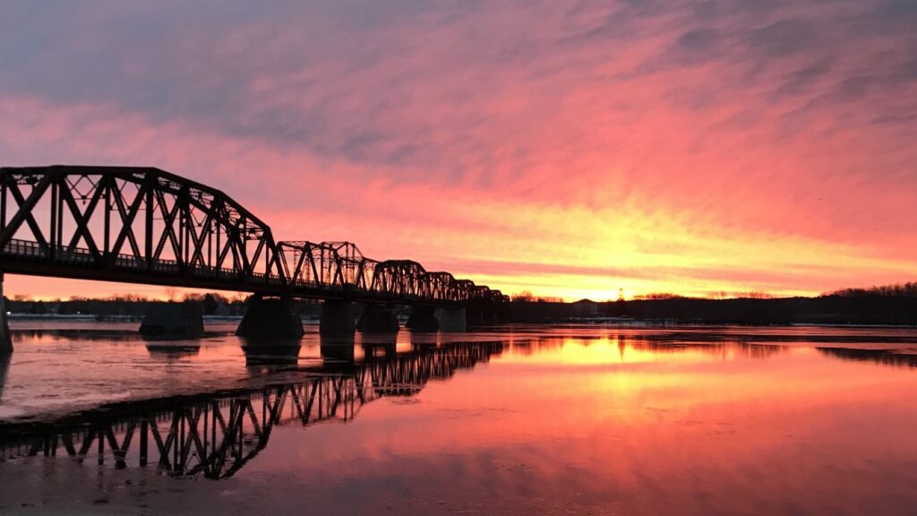 The Bill Thorpe Pedestrian Bridge, which has large X designs on it, and it reflects in the water of the Wolastoq river. The sunset is pink/yellow/blue, which reflects on the water as well.