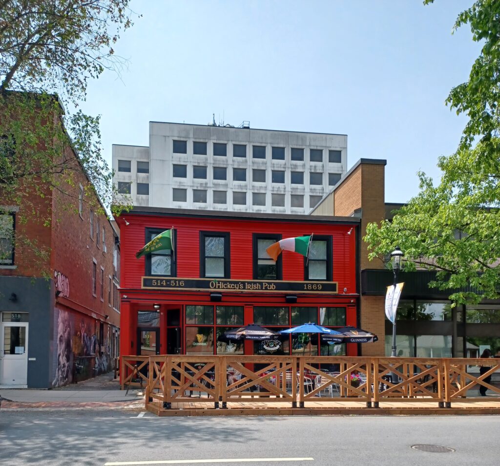 A bright red building that says "O'Hickey's Irish Pub" with two flags at the top (the one on the right is the Irish flag). There's a patio with table and sunshades in front of the building.